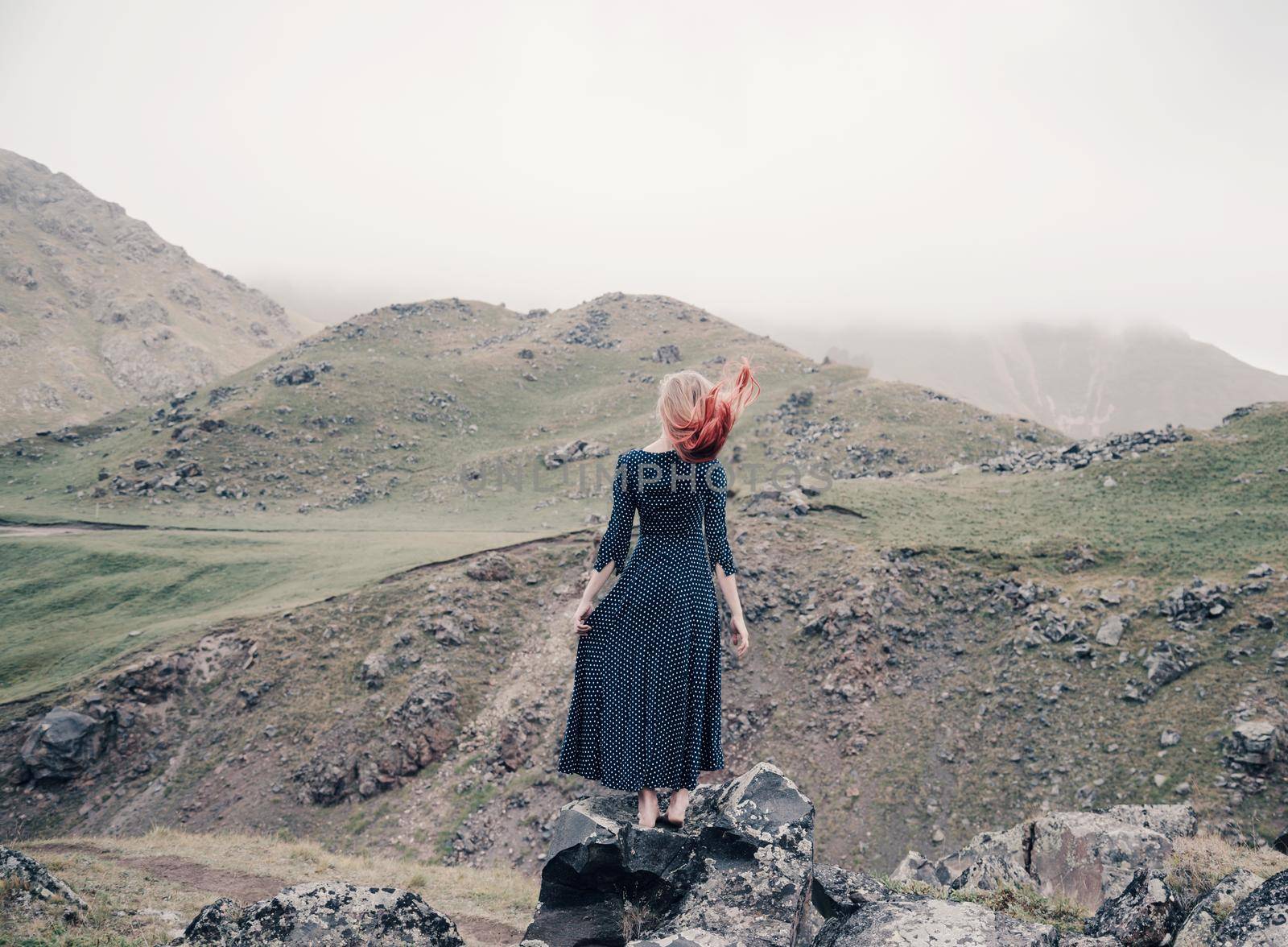 Young woman wearing in long dress standing on rocky stone in the mountains, rear view.