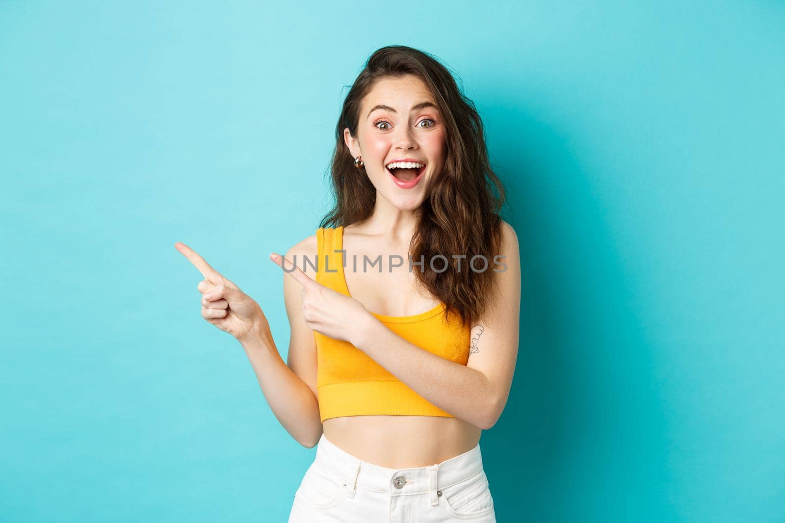 Portrait of happy young woman smiling amused, showing awesome promo deal, pointing fingers left at copy space and looking fascinated at camera, standing in summer clothes against blue background by Benzoix