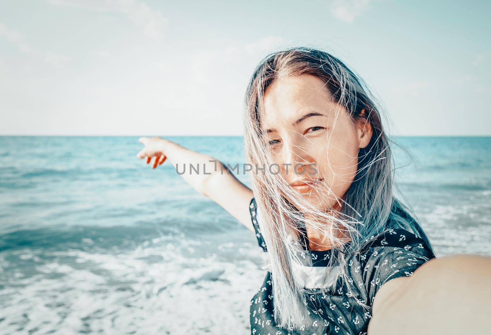 Happy young woman looking at camera on beach and pointing at the sea, point of view.