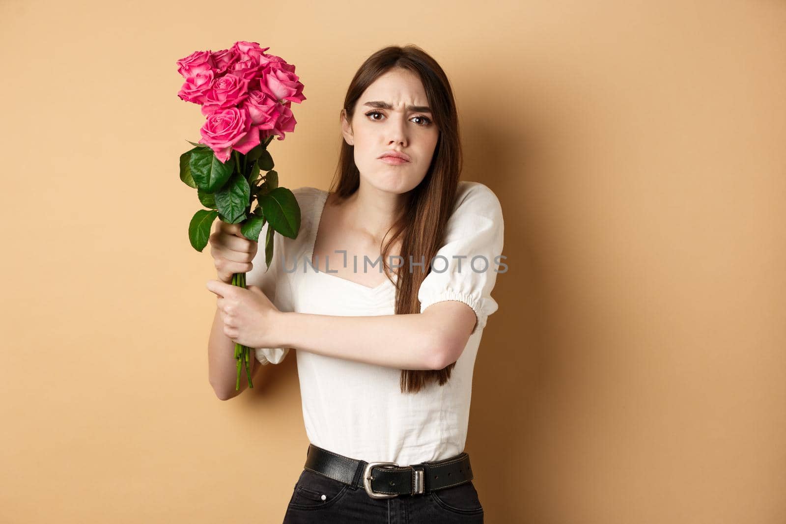 Happy valentines day. Angry girl hitting someone with flowers, swinging bouquet of roses at boyfriend, fighting with lover, standing on beige background by Benzoix
