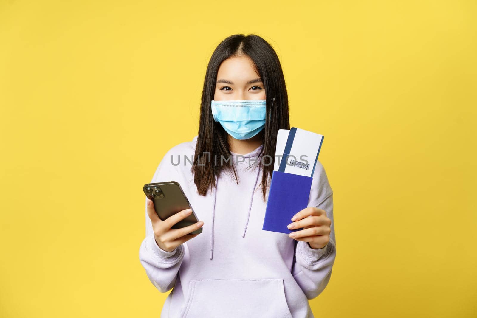 Asian tourist, girl in face mask, holding mobile phone, showing passport and flight tickets, booking hotel for vacation, going abroad, yellow background by Benzoix