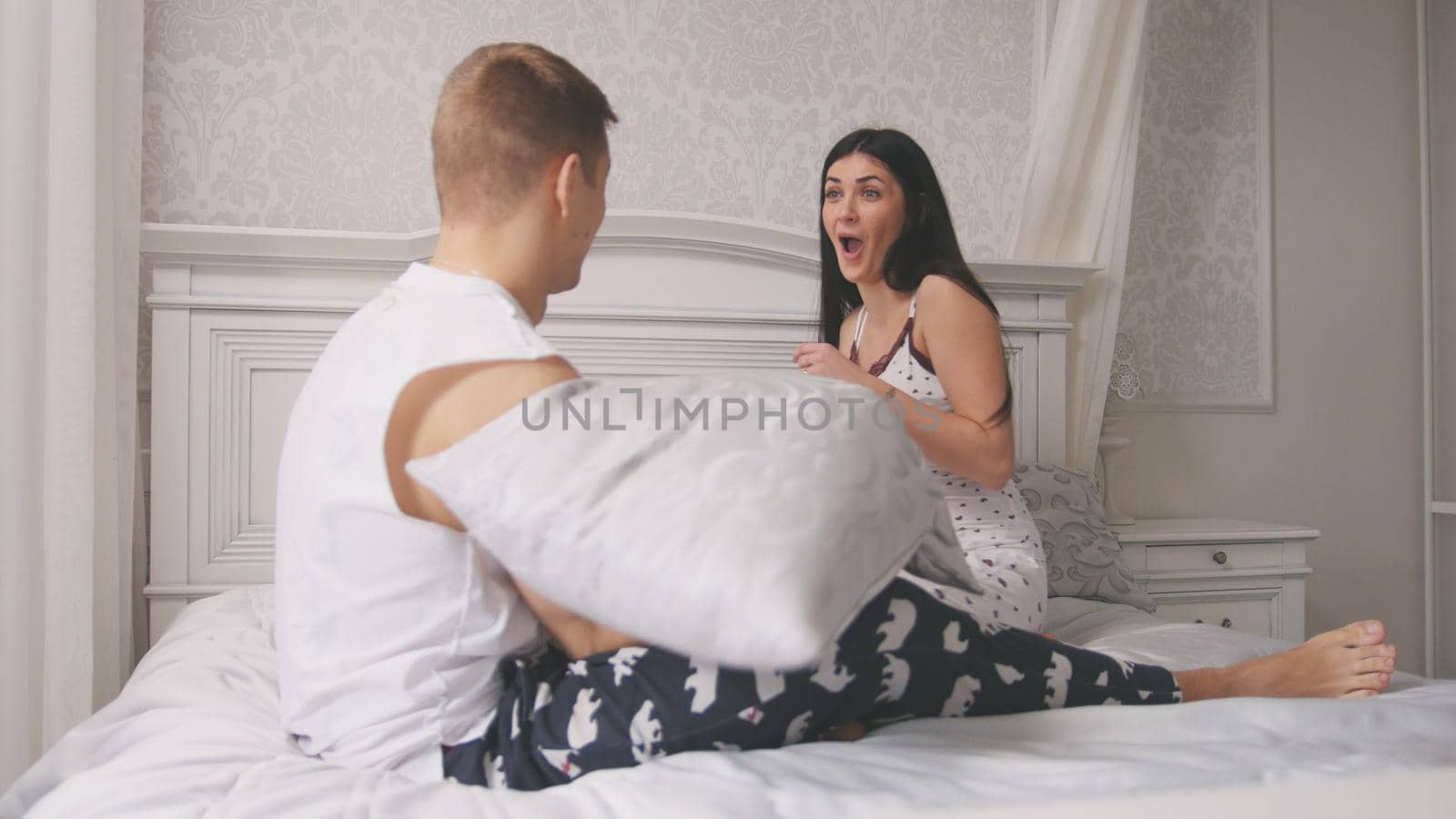 Happy cheerful loving couple having a pillow fight in bed, young attractive guy and girl sitting in pajamas, people's attitudes, close up