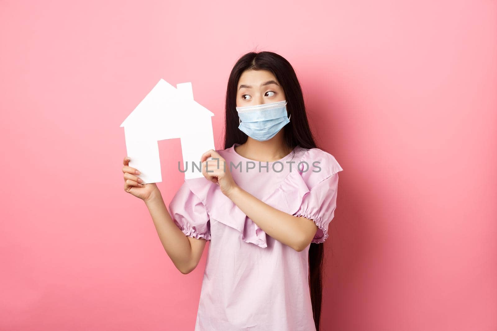 Real estate and pandemic concept. Cute asian woman in medical mask showing paper house cutout, searching for property. standing against pink background by Benzoix