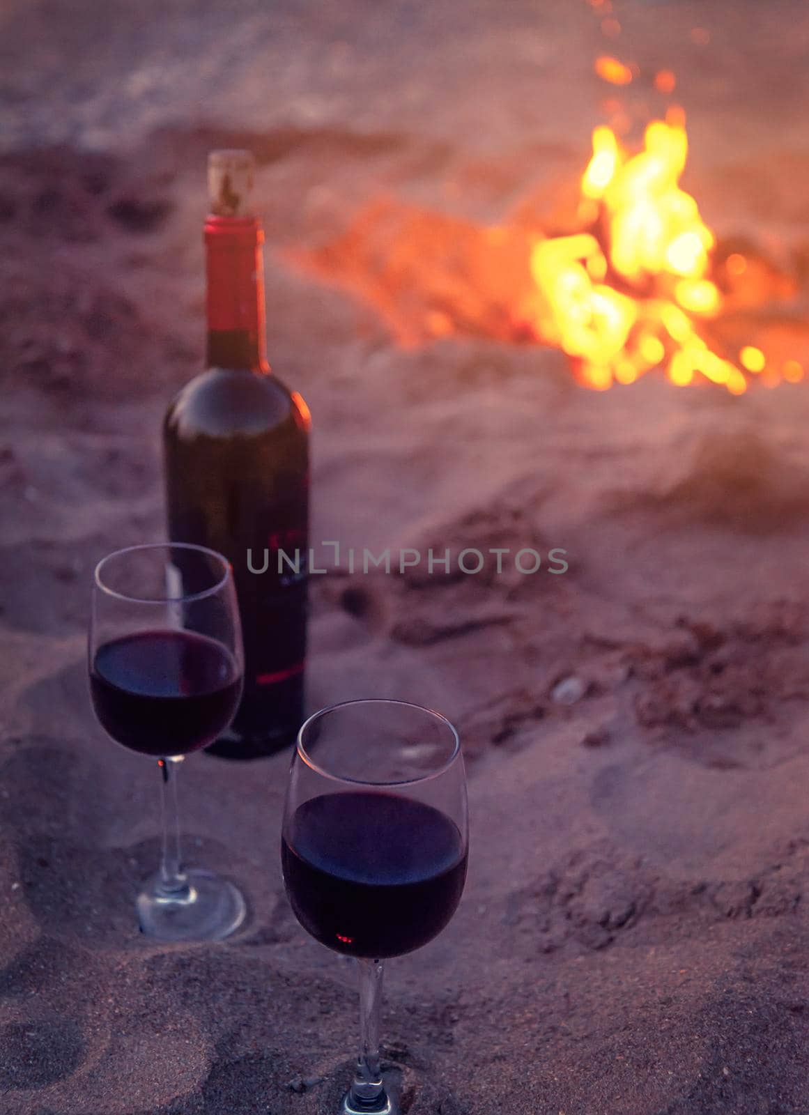 Bottle and two glasses of red wine on sand beach near campfire, concept of romantic date outdoor.