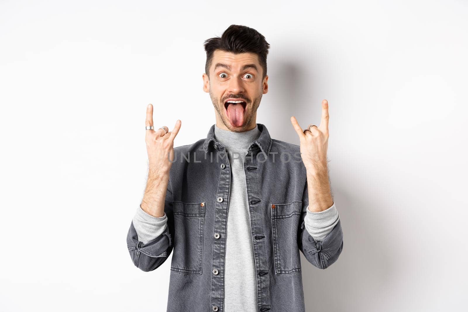 Excited funny guy showing tongue and heavy metal horns sign, enjoying party or event, having fun, standing on white background.