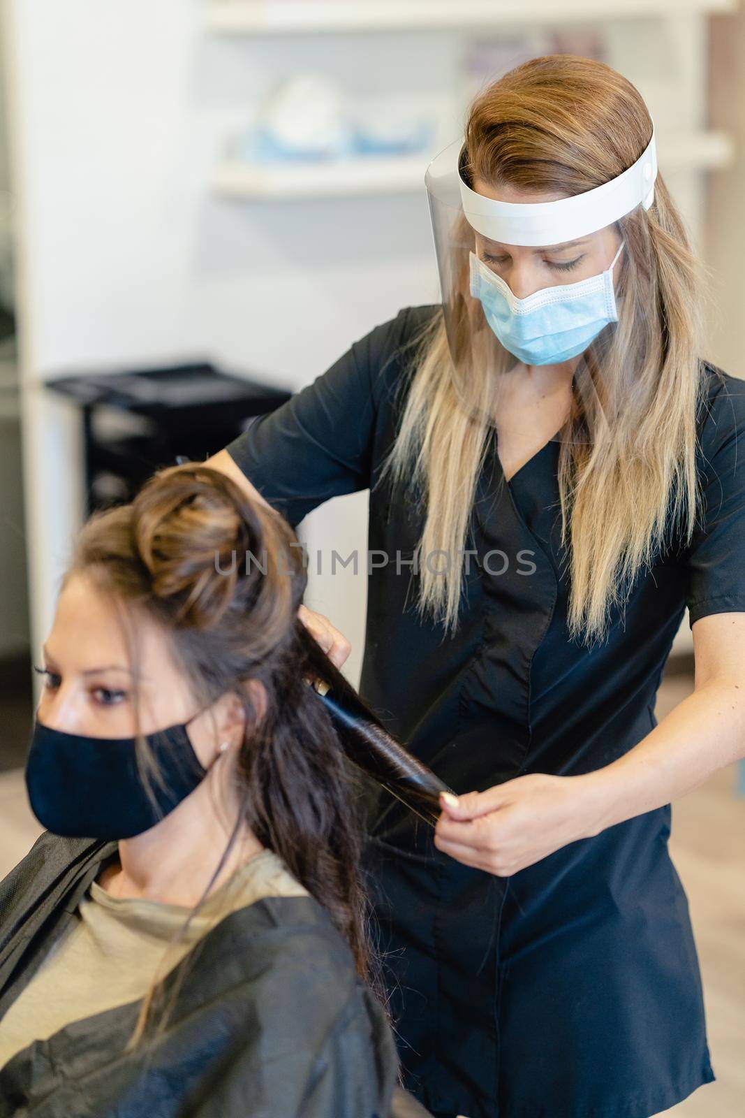 Hairdresser, protected by a mask, combing her client's hair with a hair iron in a salon. by javiindy