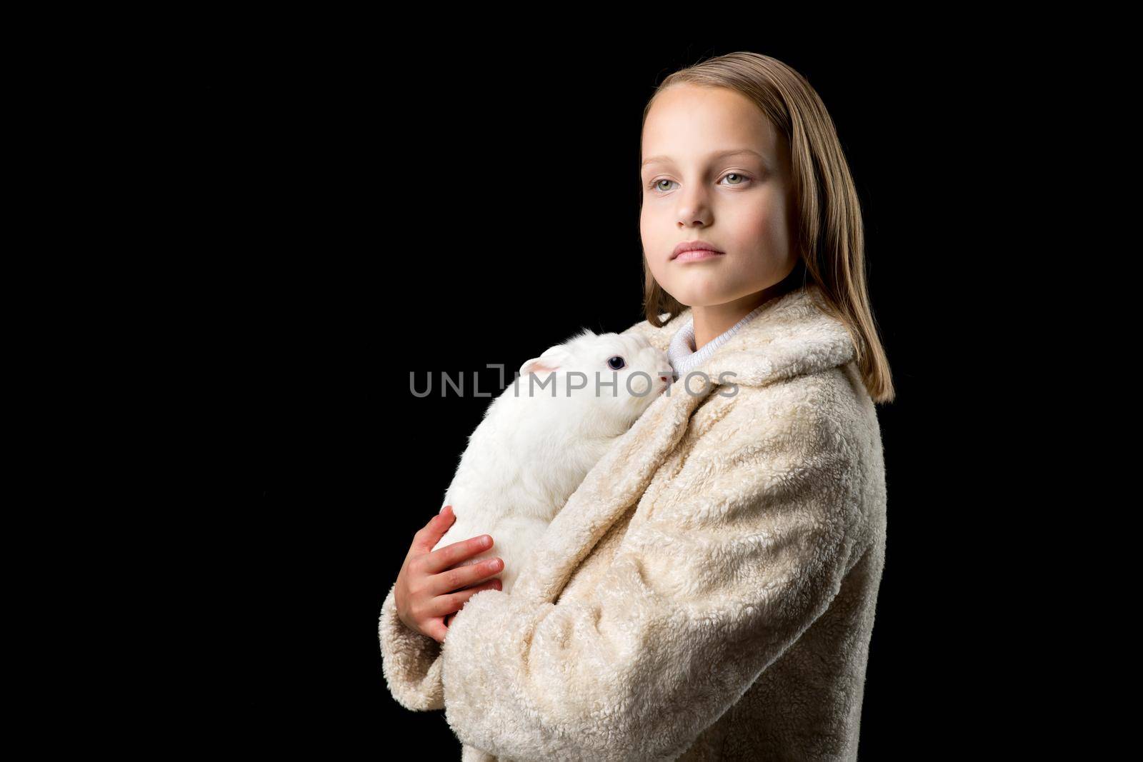 Pretty girl hugging white rabbit. Stylish beautiful girl in beige fur coat holding cute bunny. Portrait of preteen child posing with pet animal in studio against black background