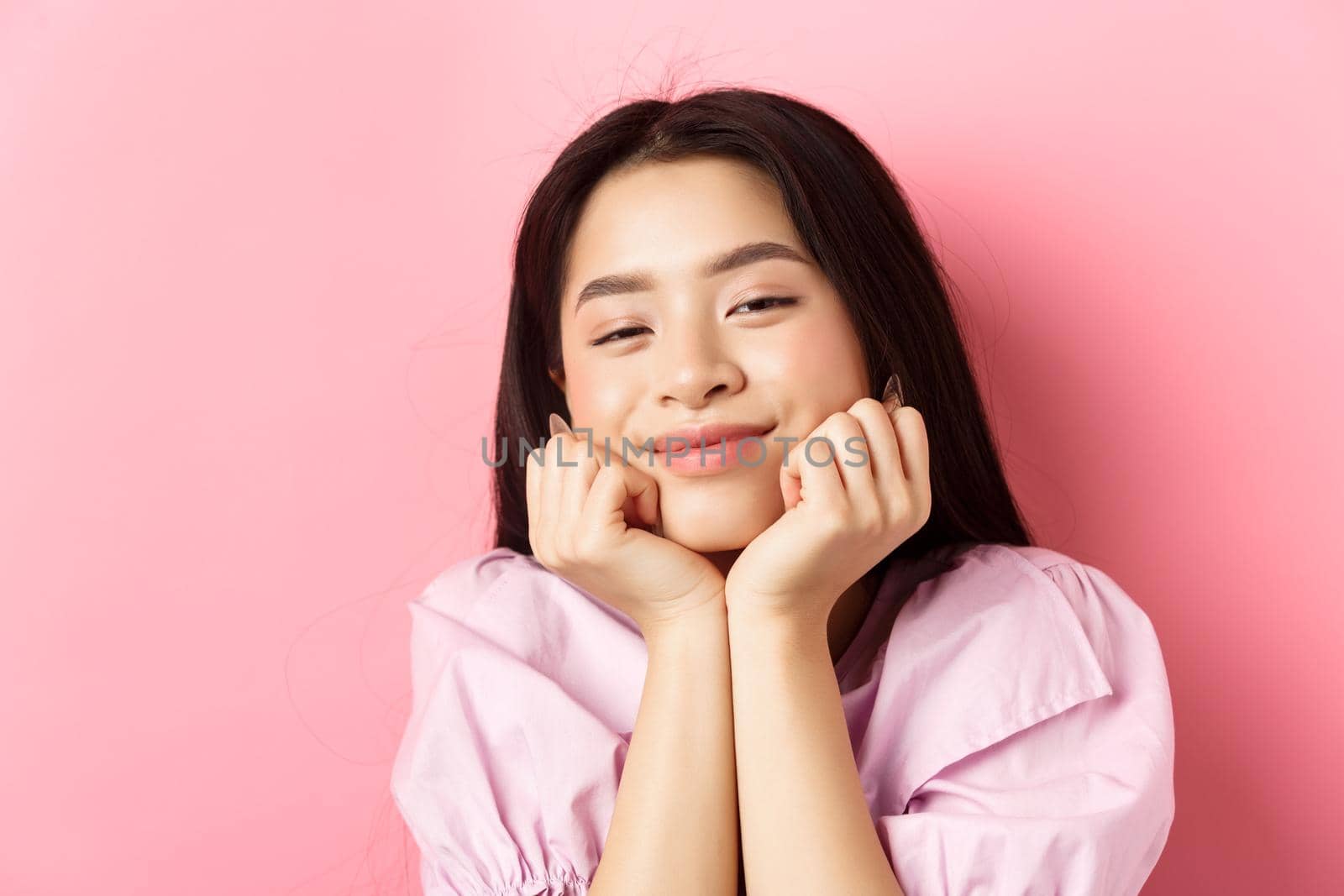 Close-up portrait of dreamy and romantic asian girl, lean face on hands and smiling, looking with admiration and happiness, standing against pink background.