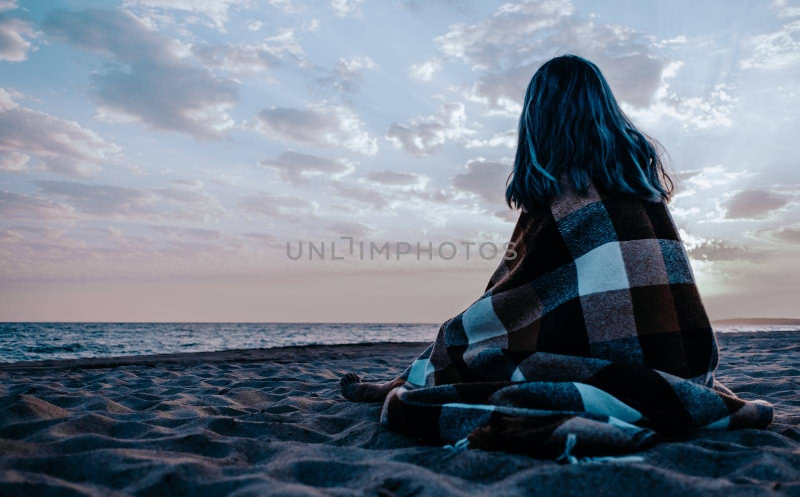 Unrecognizable young woman wrapped in plaid relaxing on sand beach near the sea in the evening, rear view.