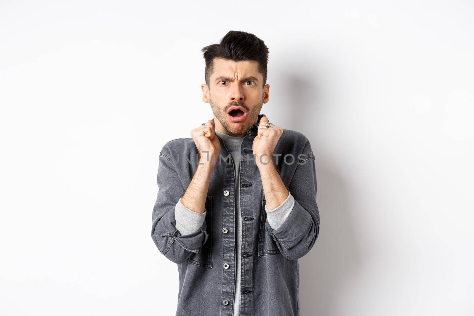Shocked young man look confused and offended, gasping and frowning at camera, hear bad news, standing on white background.
