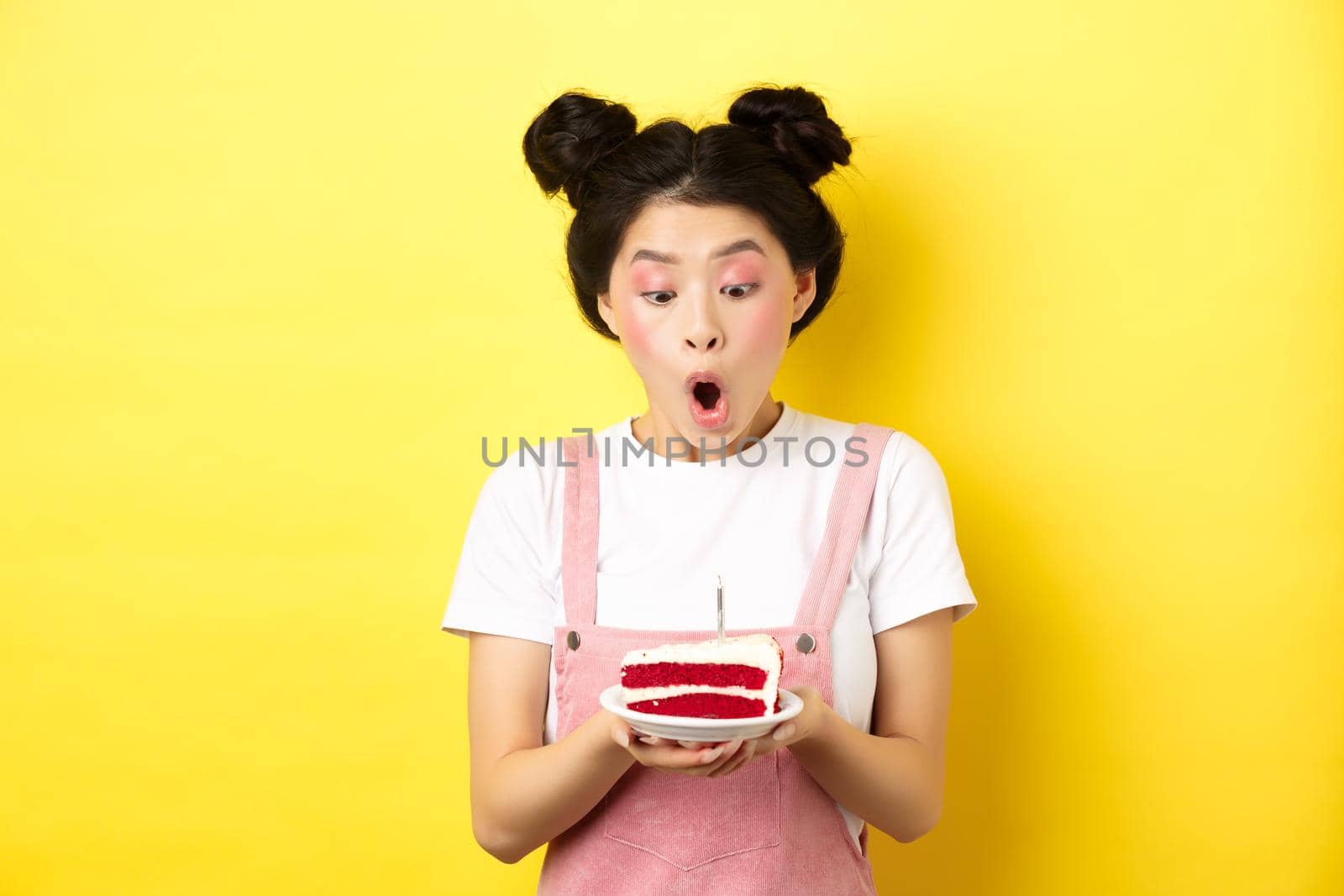 Cute asian birthday girl with bright makeup, blowing candle on cake, making wish, standing on yellow background.