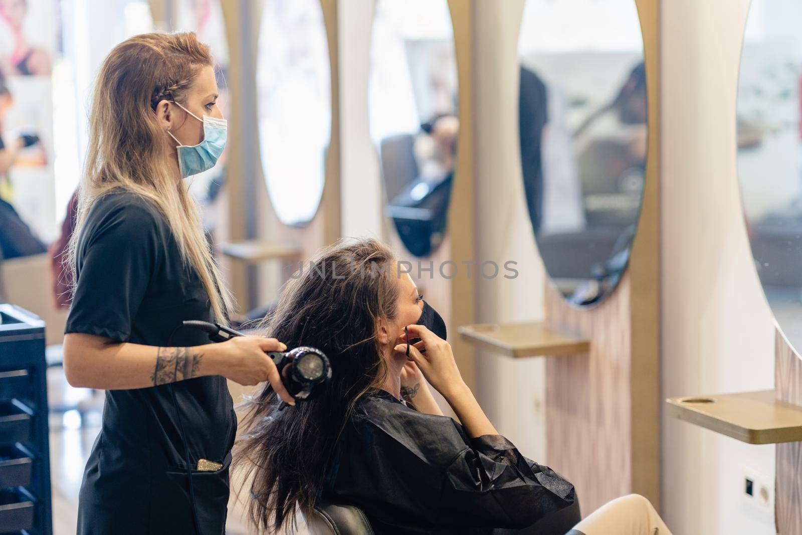 Female hairdresser drying her client's hair with a hairdryer, wearing protective masks in a beauty centre. Business and beauty concepts