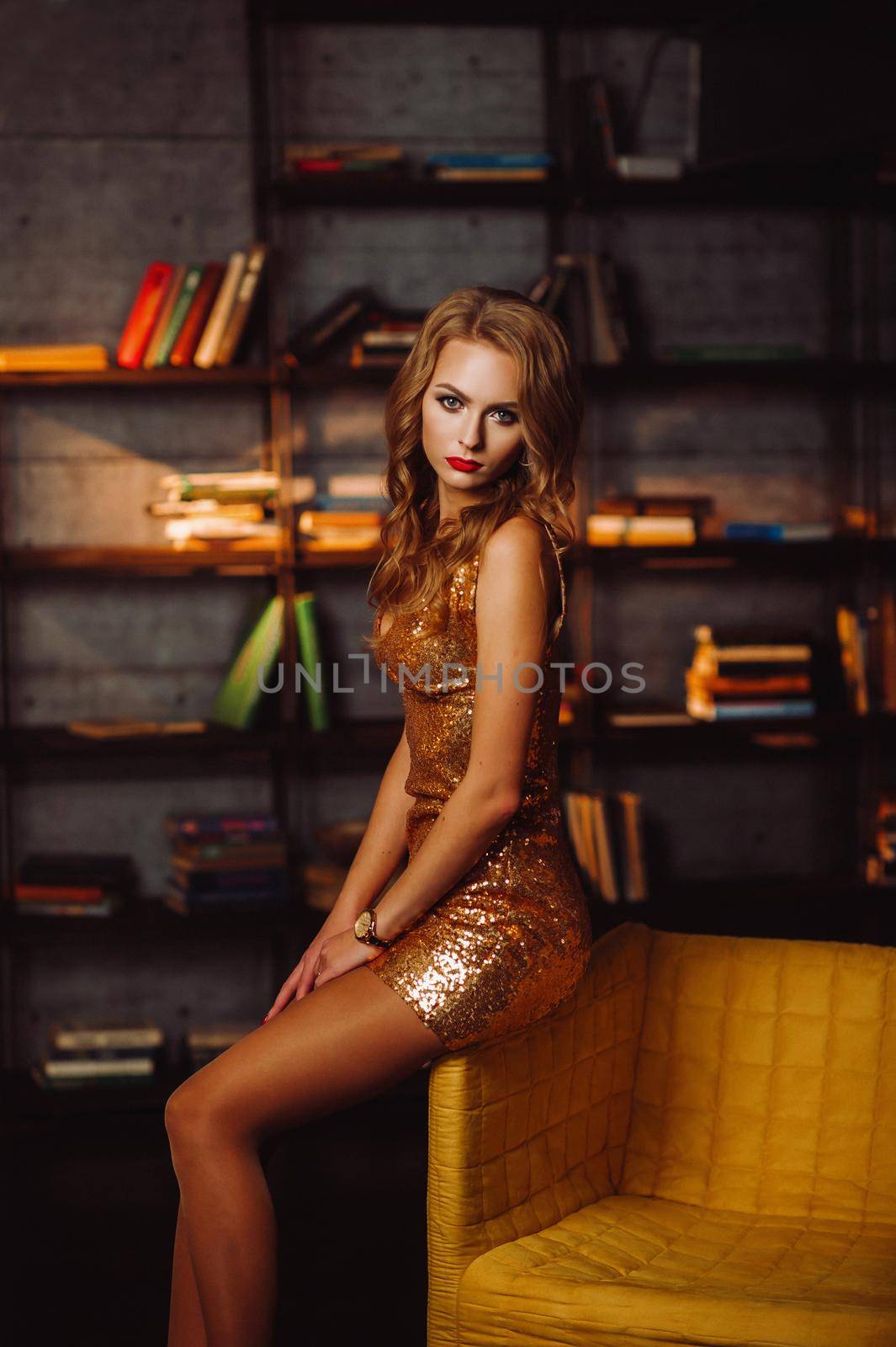 Sexy girl in a gold dress on the background of books in the library.Model with long hair and red lipstick in the interior by Lobachad