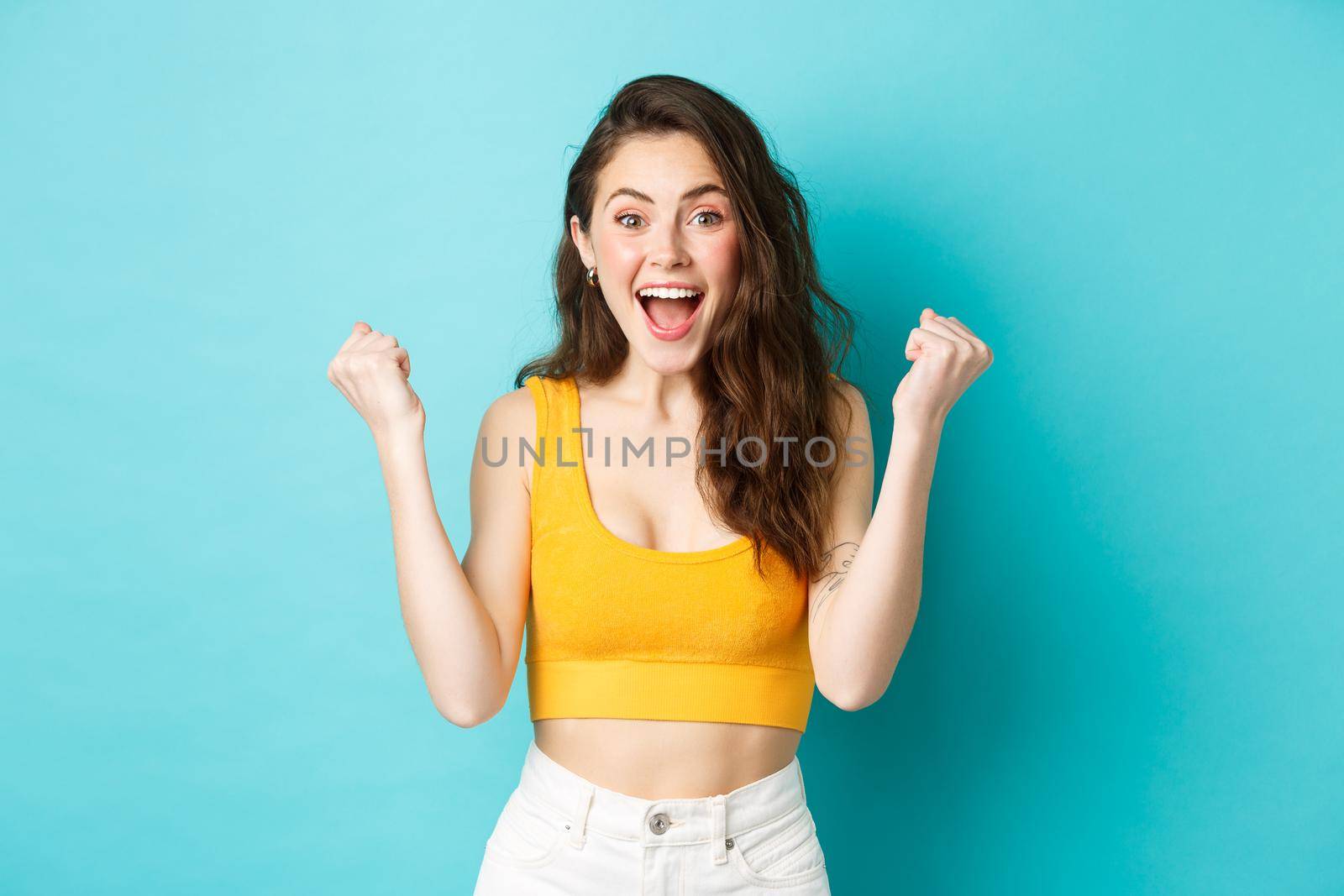 Summer holidays and emotions concept. Excited young woman winning and screaming from joy, achieve goal, celebrating victory, making fist pumps, standing over blue background.