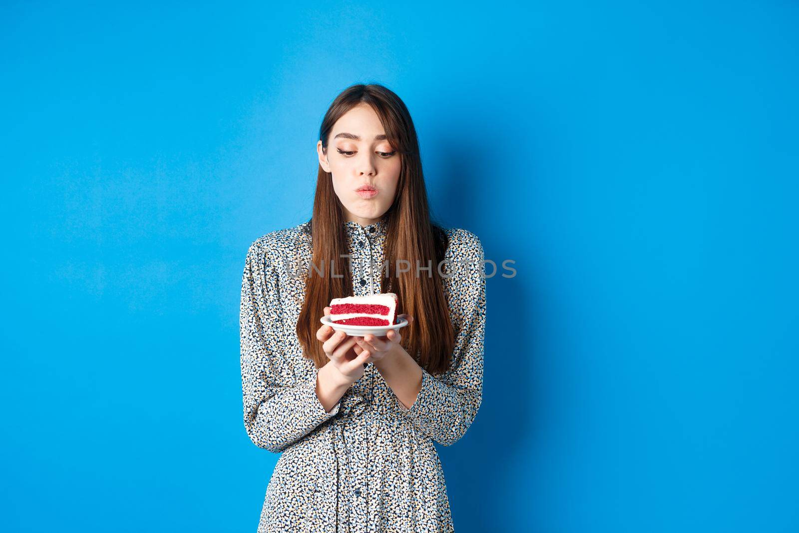 Cute birthday girl celebrating, blowing candle on cake and making wish, standing on blue background. Party and holidays concept.