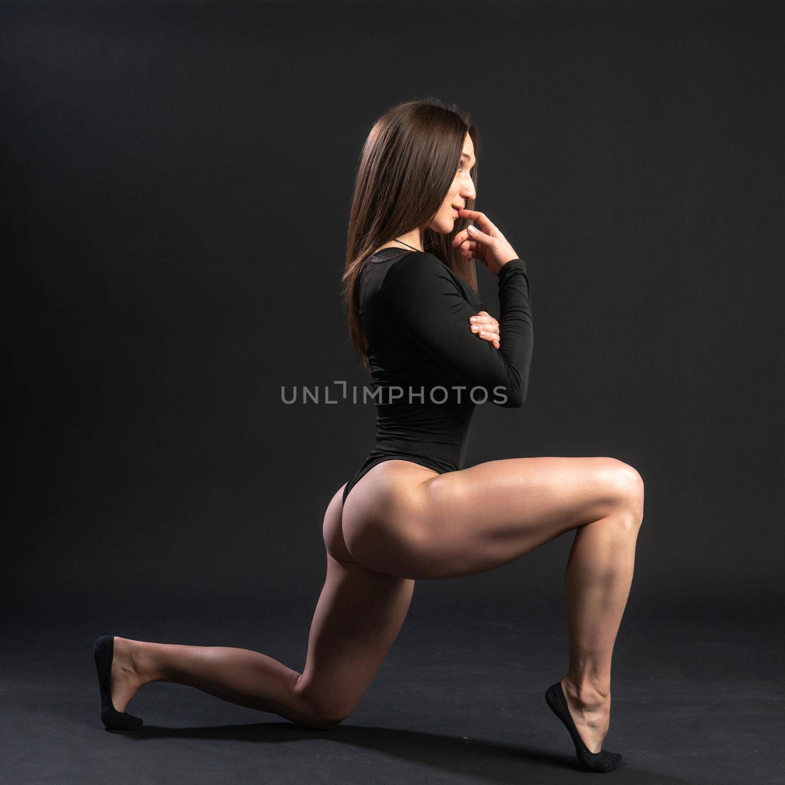 The girl is beautiful in a black swimsuit bodybuilder stands on his knee and erotically touches the lips. Bare feet on a dark background
