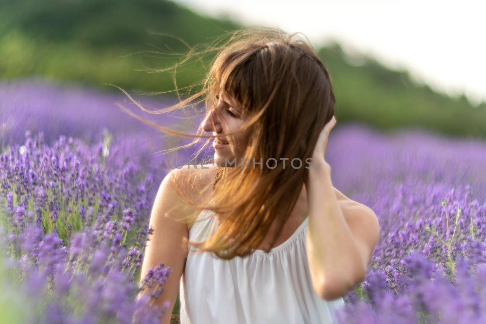 Close up portrait of happy young brunette woman in white dress on blooming fragrant lavender fields with endless rows. Warm sunset light. Bushes of lavender purple aromatic flowers on lavender fields. by panophotograph