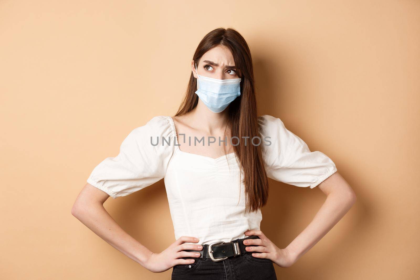 Covid-19 and lifestyle concept. Suspicious woman in medical mask frowning, looking aside at logo with doubtful face, standing on beige background.