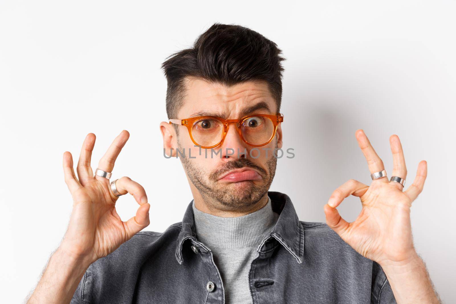 Not bad. Funny young guy in glasses pucker lips, showing okay sign and look silly at camera, standing on white background.