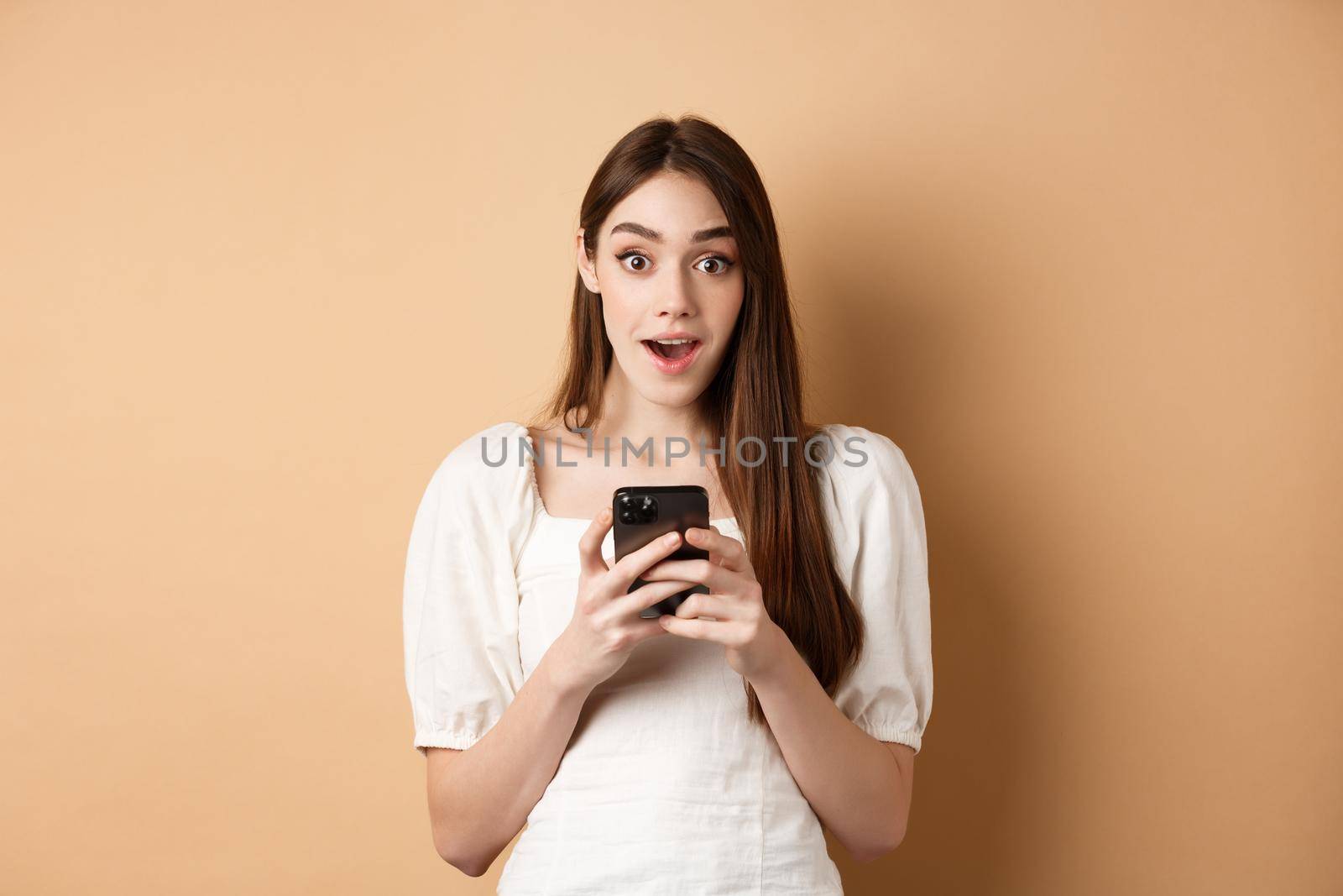 Exciting news on phone. Amazed young woman say wow after reading online announcement on smartphone, standing on beige background.