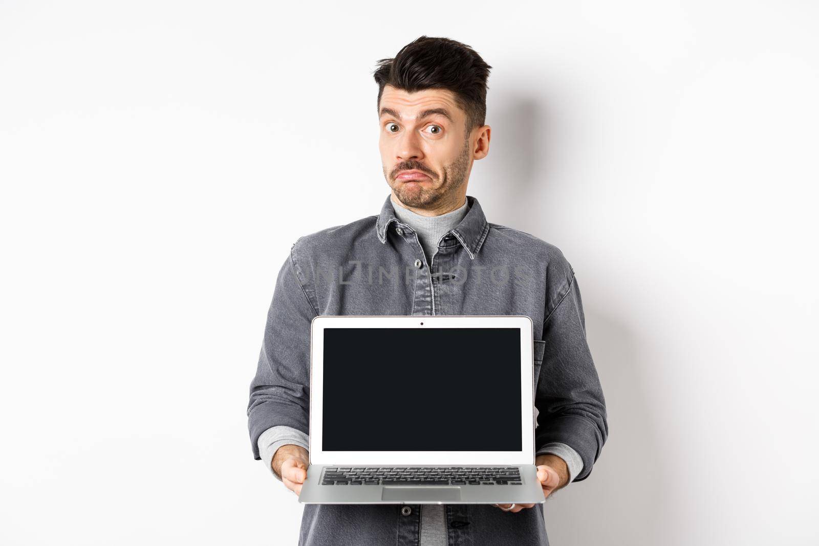 Indecisive young man pouting and shrugging while showing empty laptop screen, standing clueless on white background.