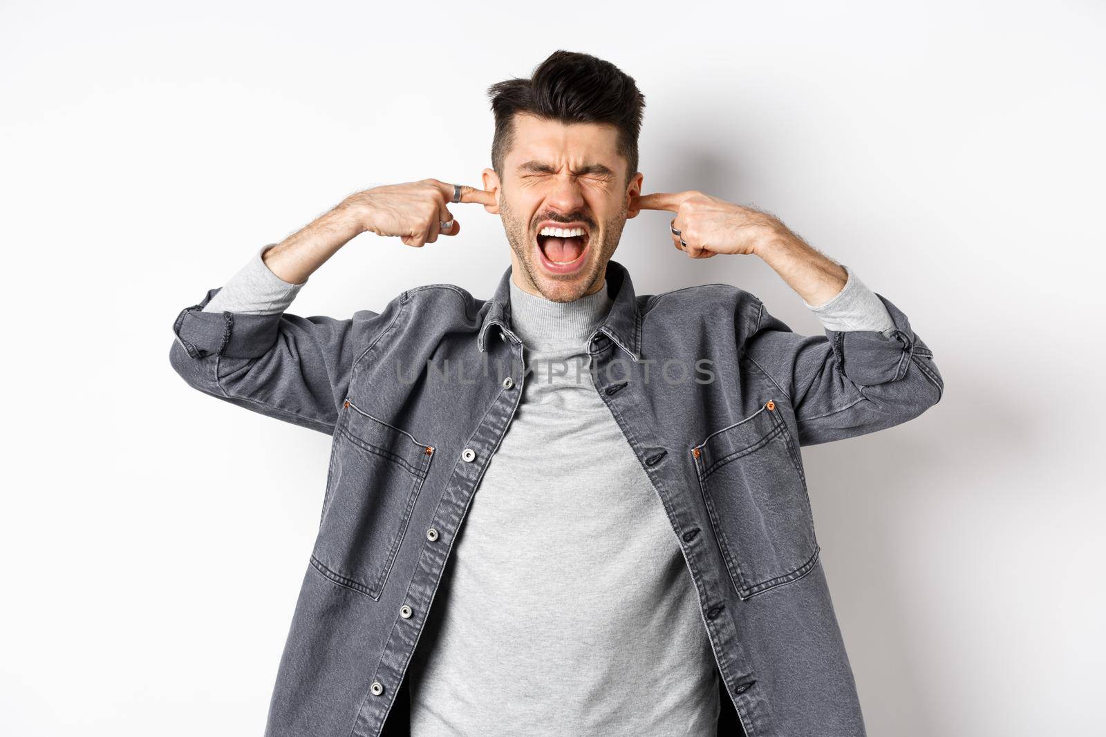 Annoyed guy shut ears and screaming, displeased by loud noise, yelling to stop noisy music, standing on white background.