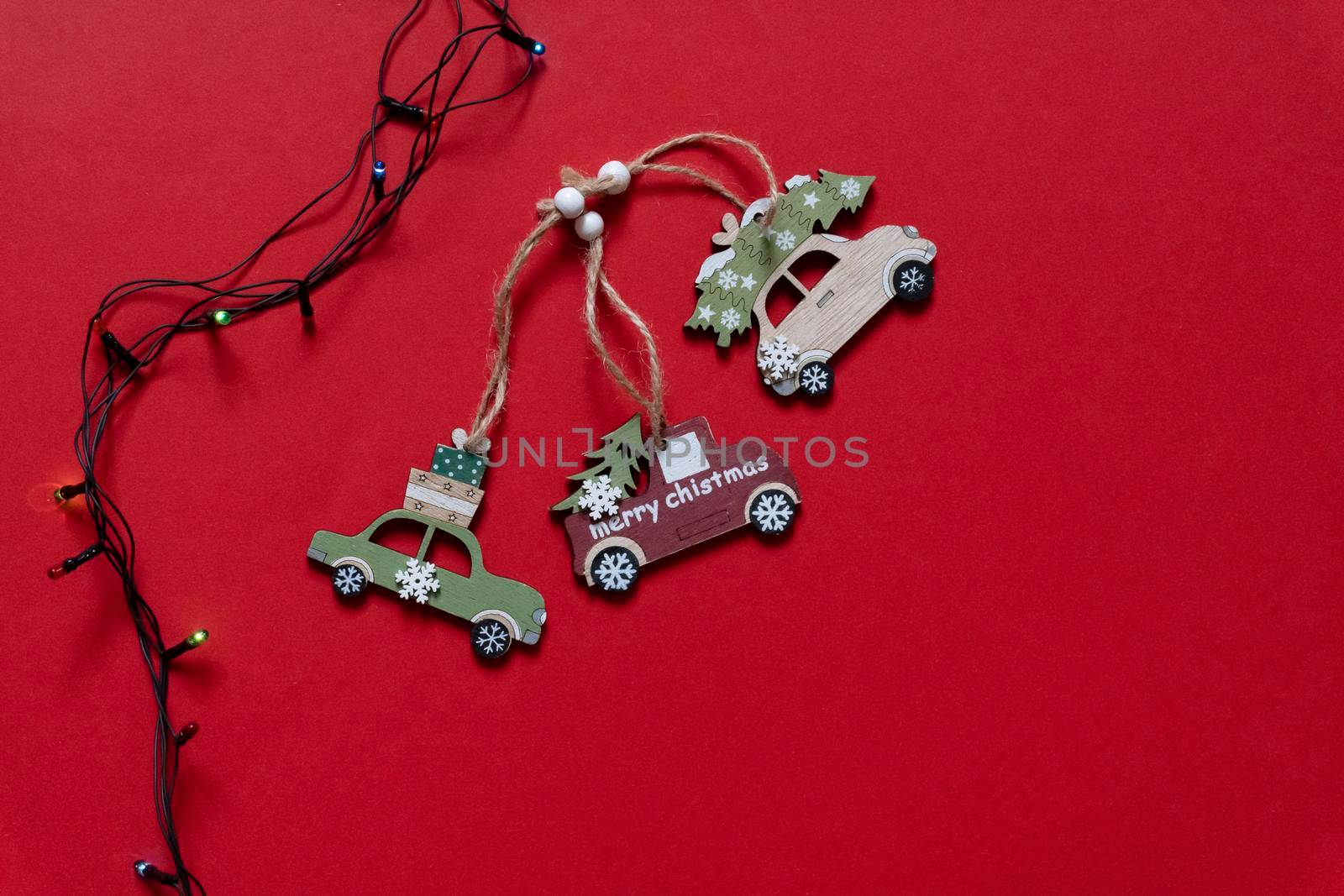 Three wooden car toys for Christmas tree near colocful light bulb on red background, top view, copy space.