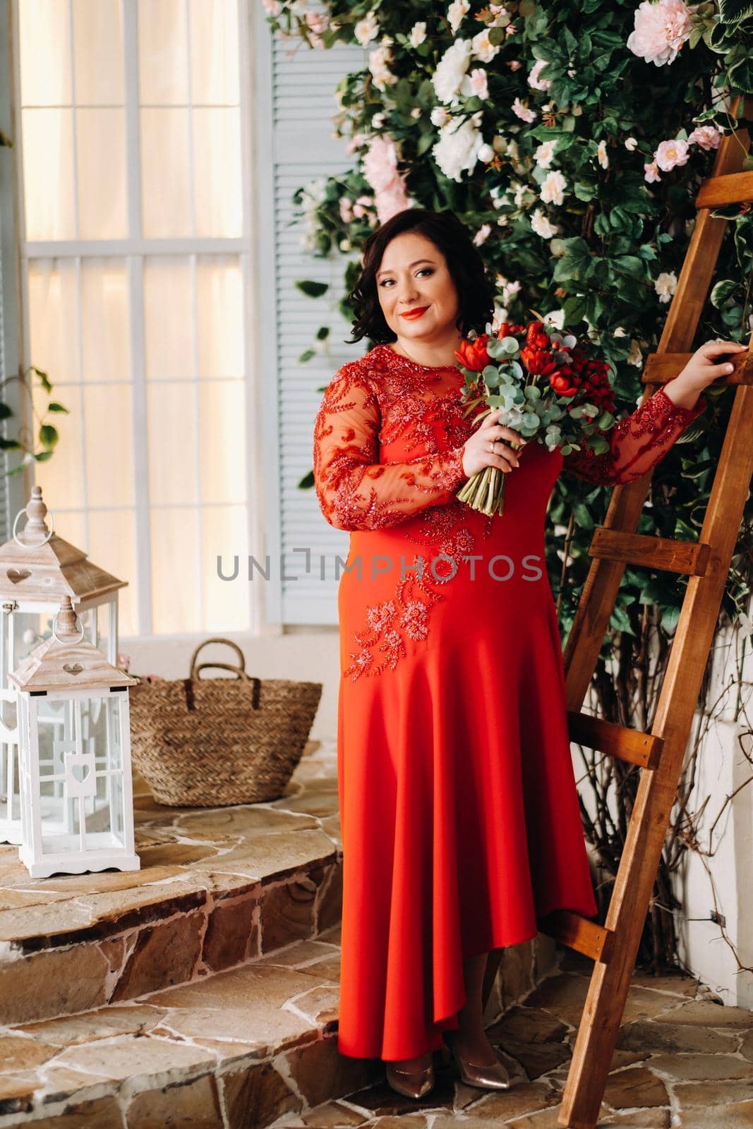 a woman in a red dress stands and holds a bouquet of red roses and strawberries in the interior