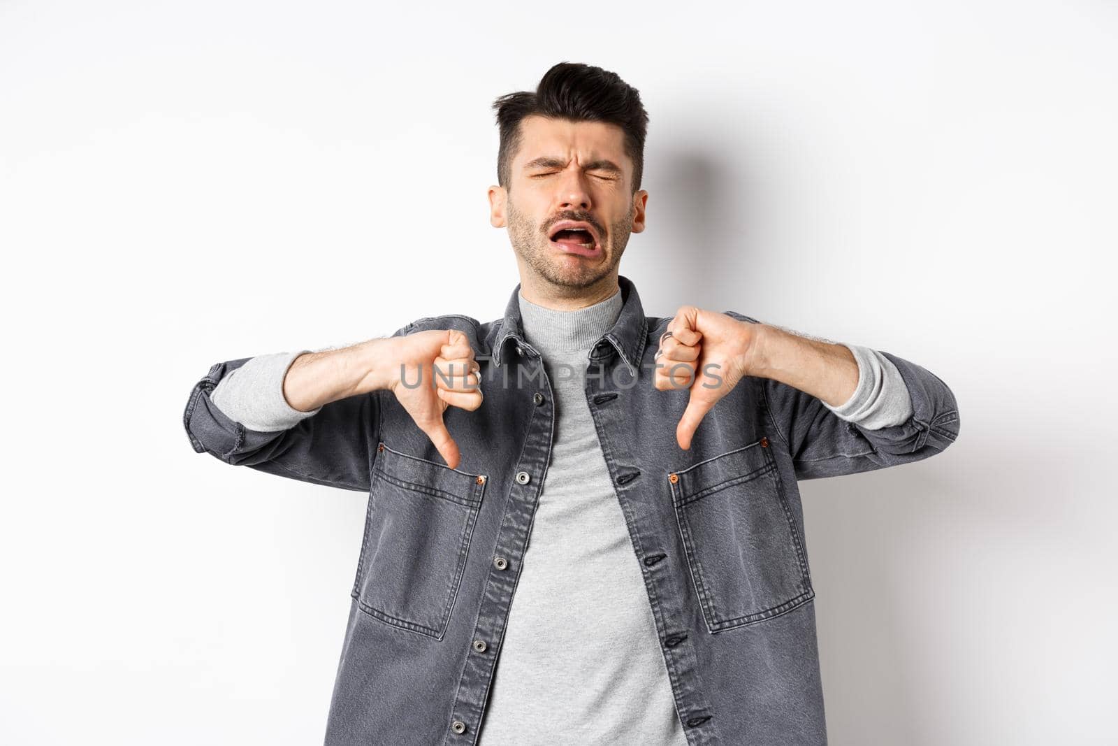 Sad man crying and showing thumbs down, whining and complainin, being upset and disappointed, standing on white background.