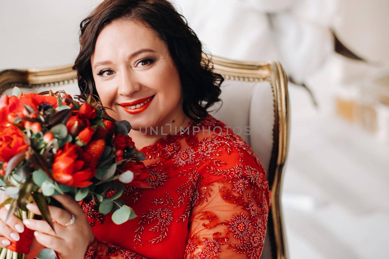 a woman in a red dress sits in a chair and holds a bouquet of red roses and strawberries in the interior