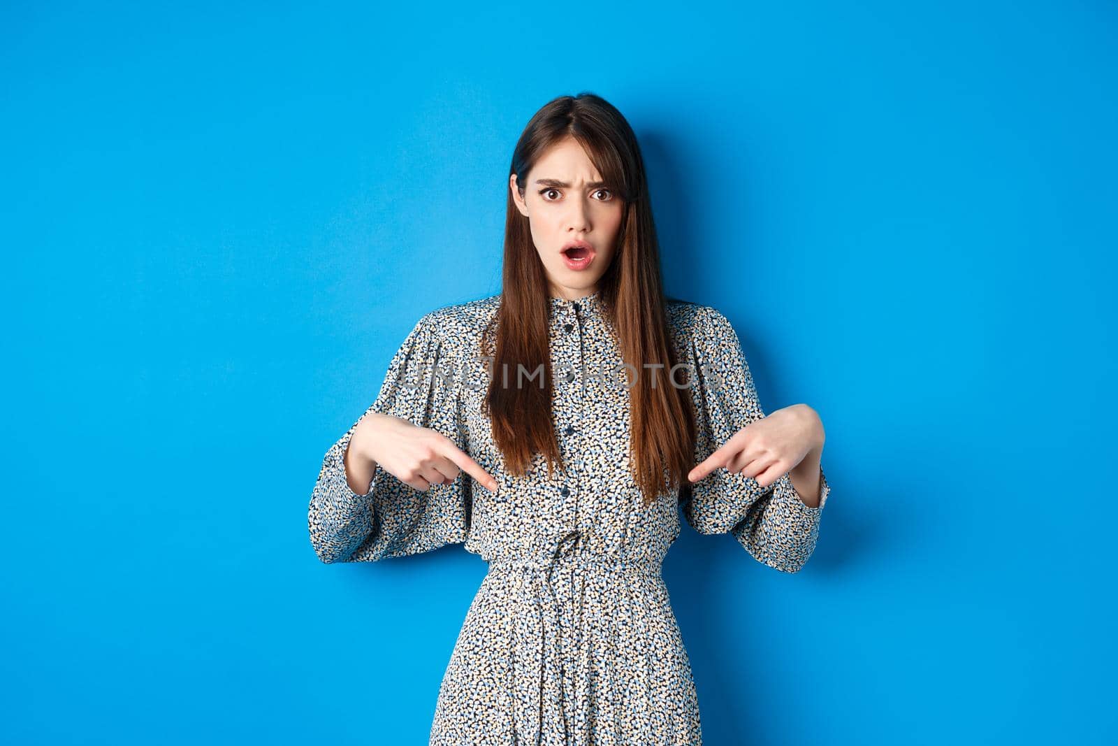 Shocked and offended young woman in dress frowning, gasping and pointing fingers down at insulting promo, standing on blue background.