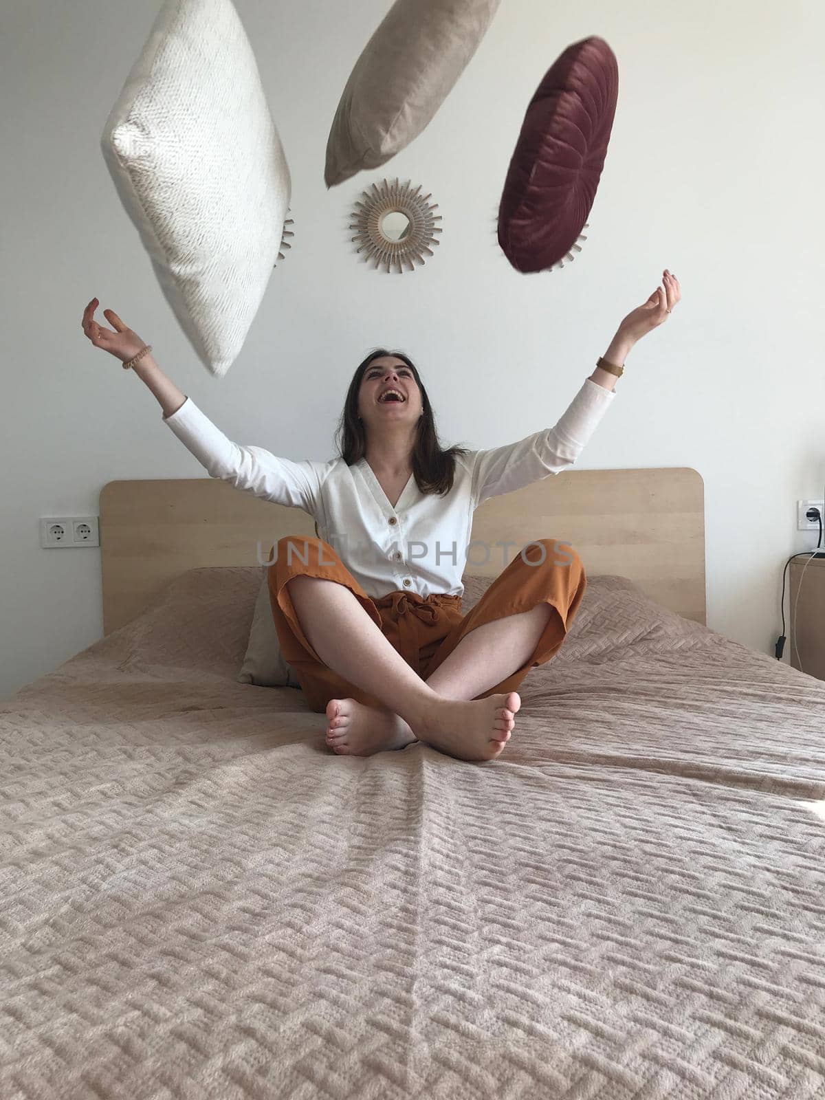 cheerful girl with loose hair sitting on the bed in her underwear tossing up the pillow and smiling.