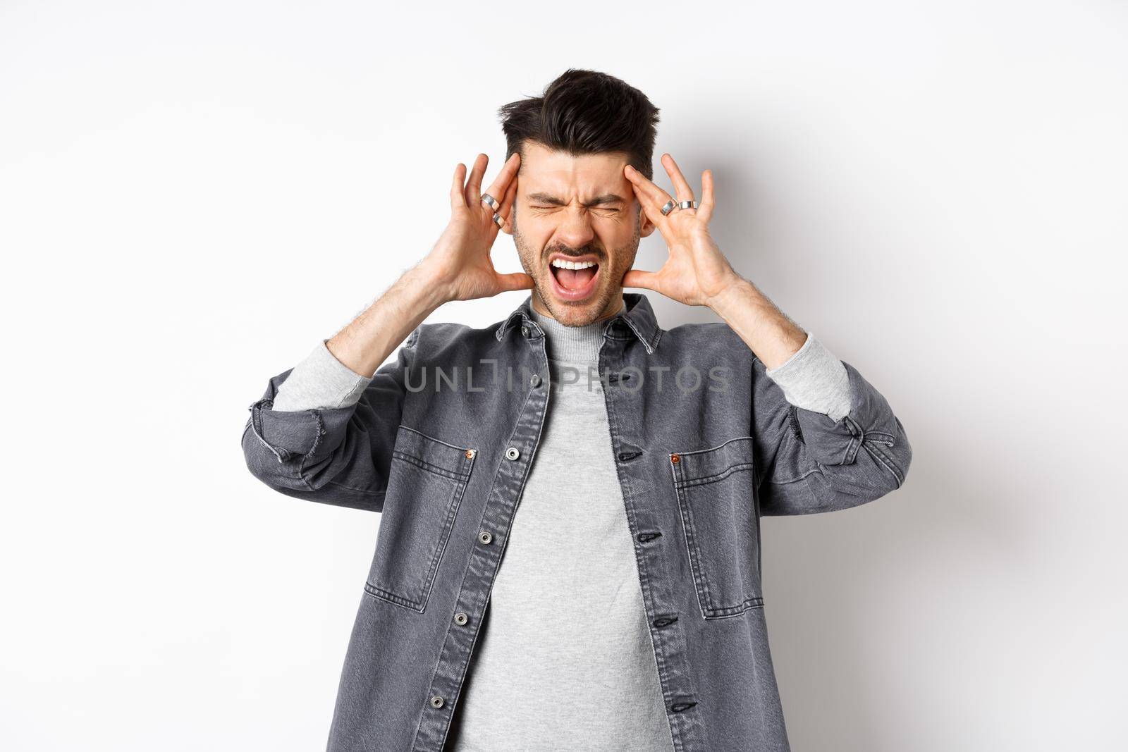 Frustrated young man shouting and touching head distressed, standing bothered by loud noise or headache, standing on white background.