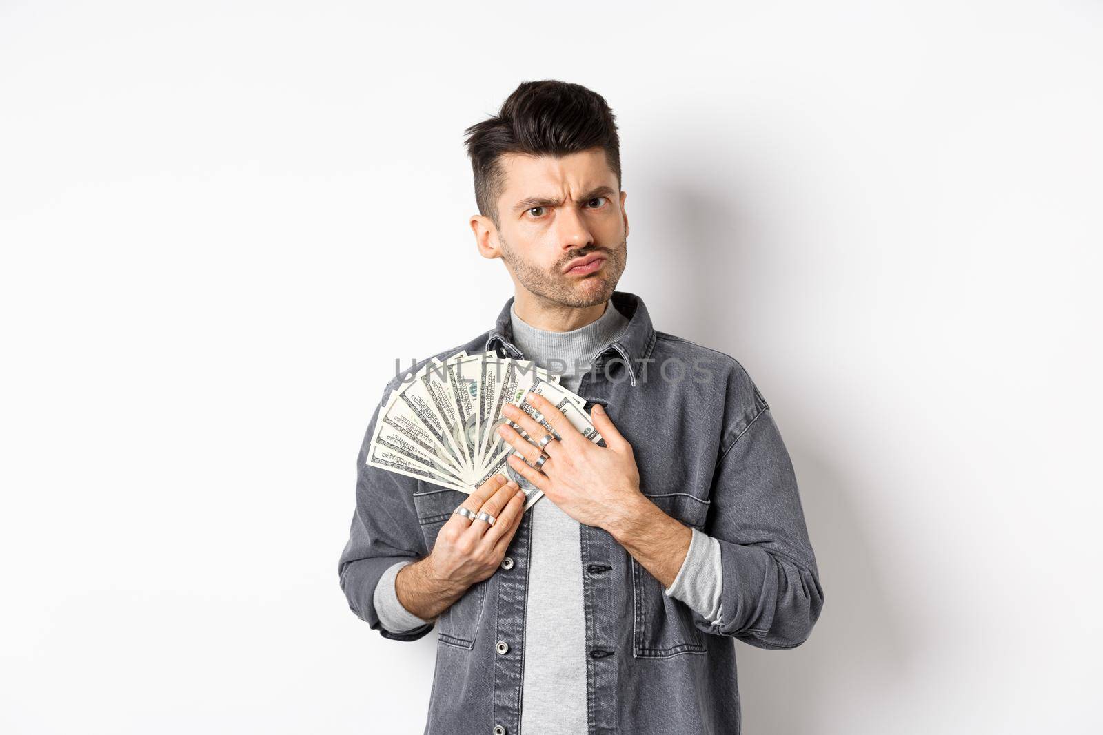 Greedy guy frowning and hugging dollar bills, unwilling to share money, standing on white background.