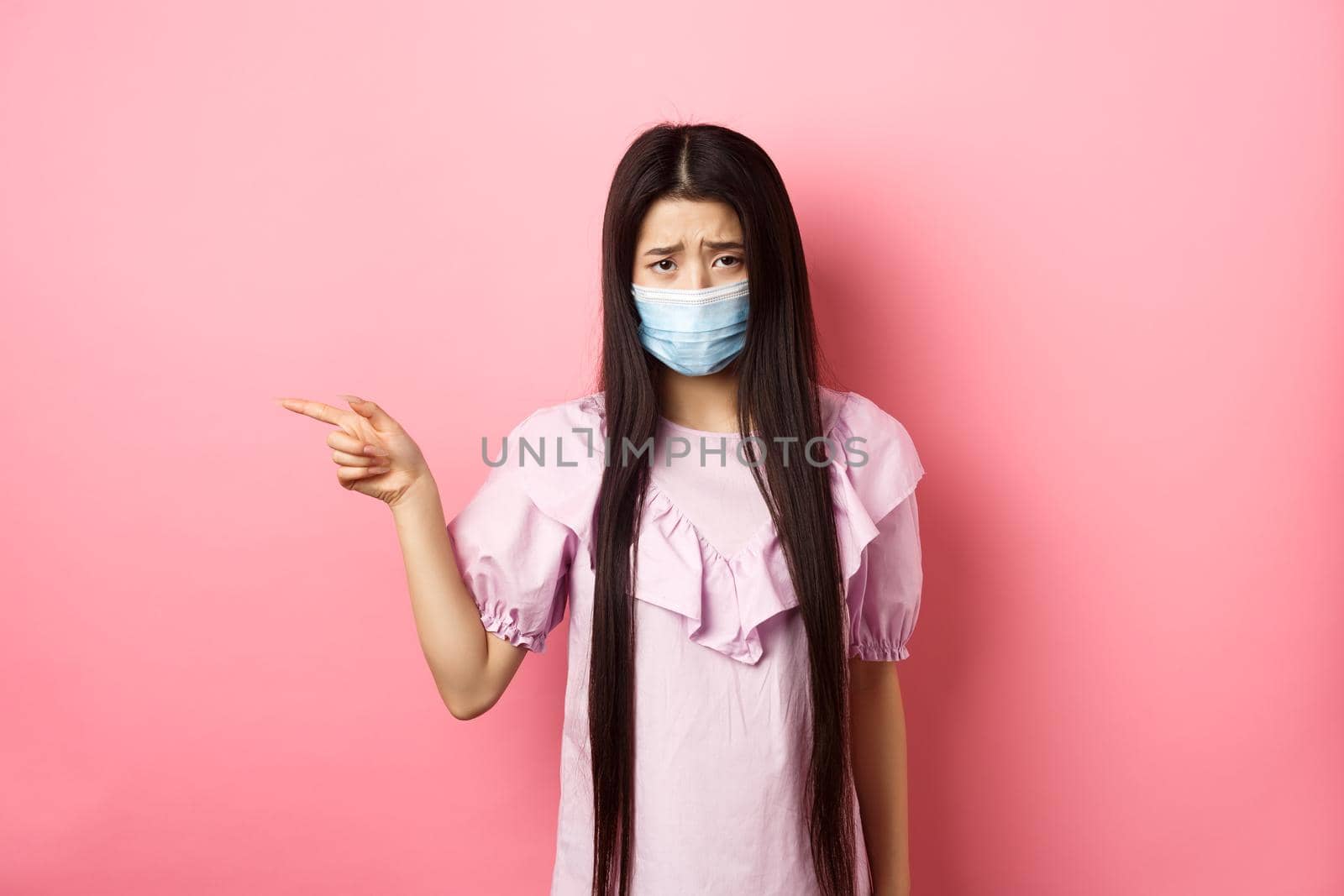 Covid-19, pandemic lifestyle concept. Sad and gloomy asian girl in medical mask pointing left, complain on unfair situation, standing disappointed against pink background.