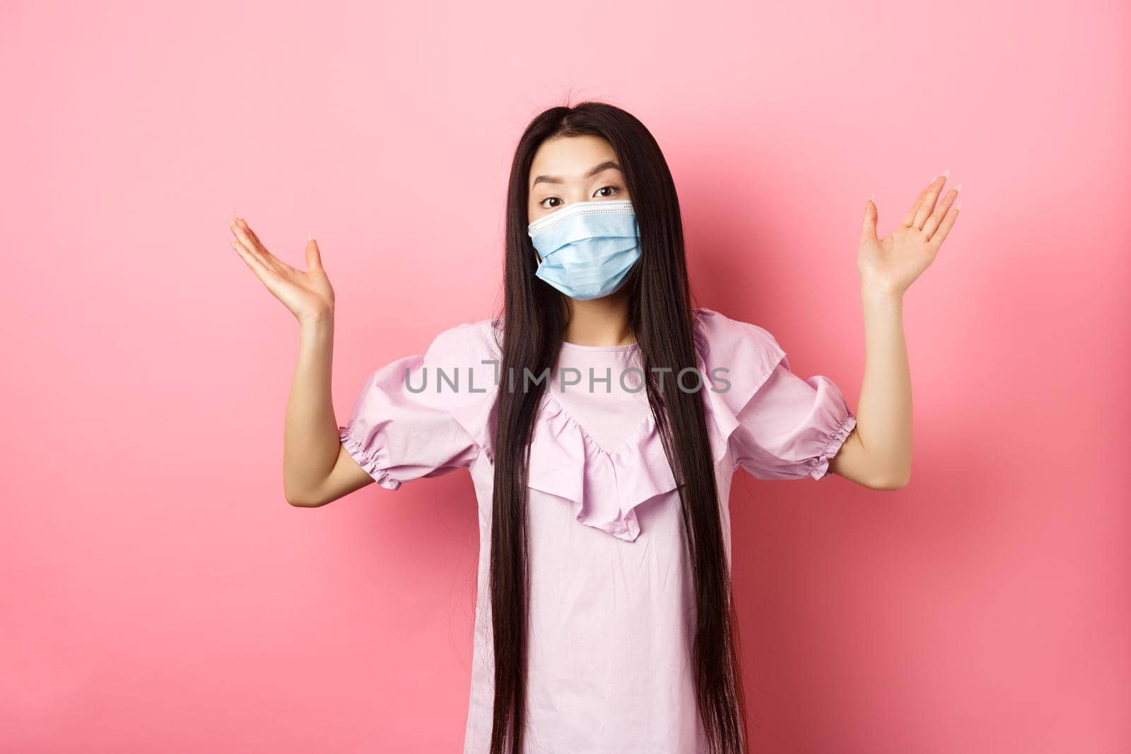 Coronavirus, quarantine and lifestyle concept. Clueless asian girl in face mask shrugging shoulders unware, know nothing, standing on pink background.