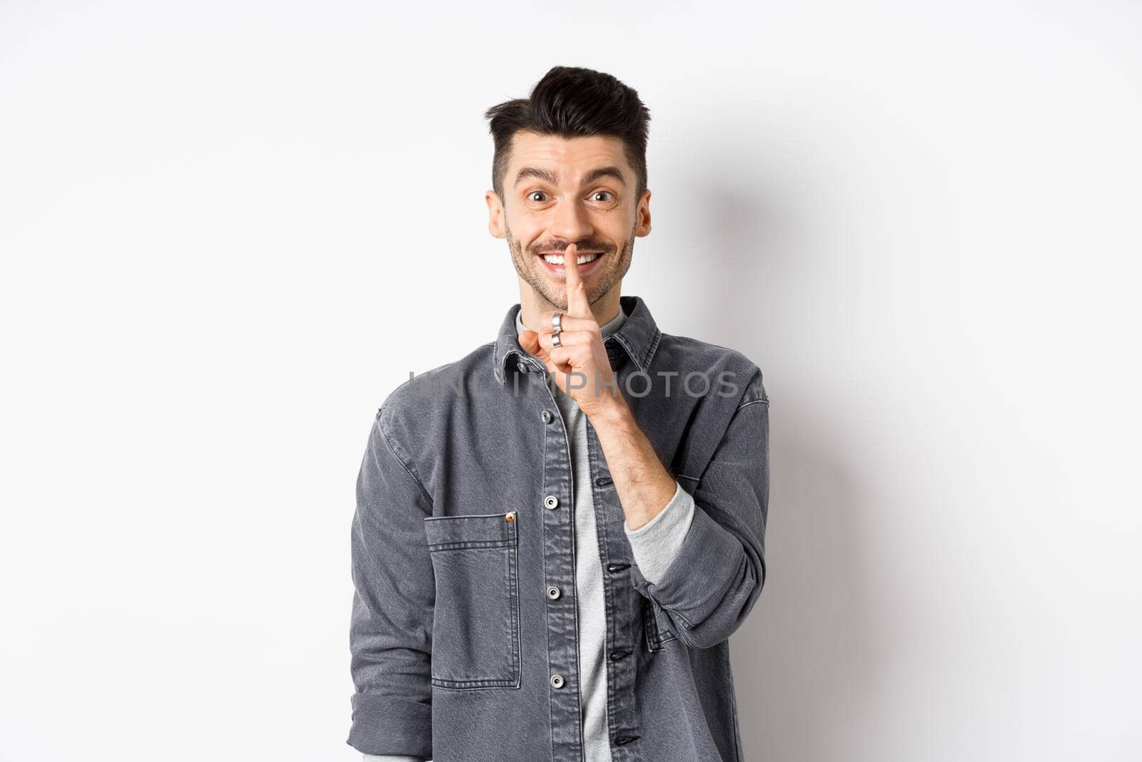 Happy guy shushing at camera, smiling with finger on lips, asking to keep quiet, telling big secret or making surprise, standing on white background.