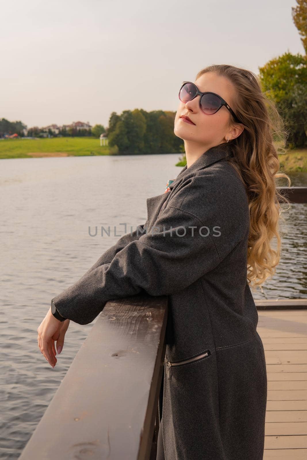 Joyful young stand near the water with a beautiful smile with white skin in a coat in the fall on the dock in the afternoon by 89167702191
