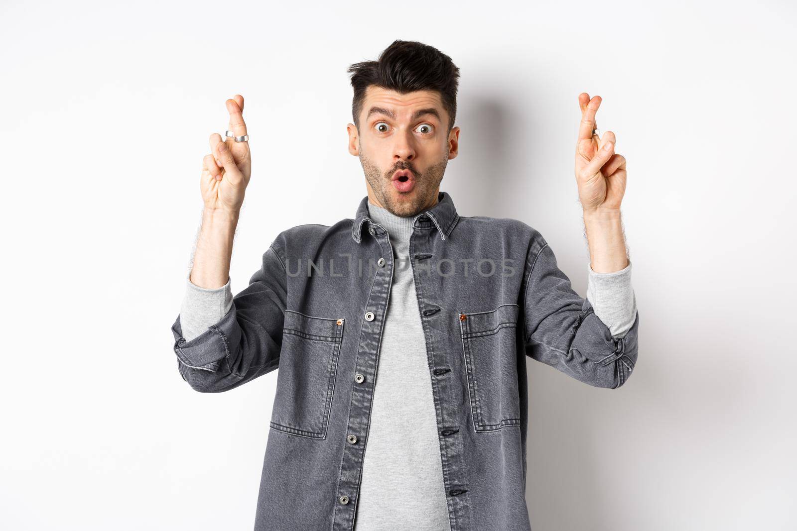 Excited young man cross fingers for good luck and gasping, holding breath as waiting important news, begging or praying, standing on white background.