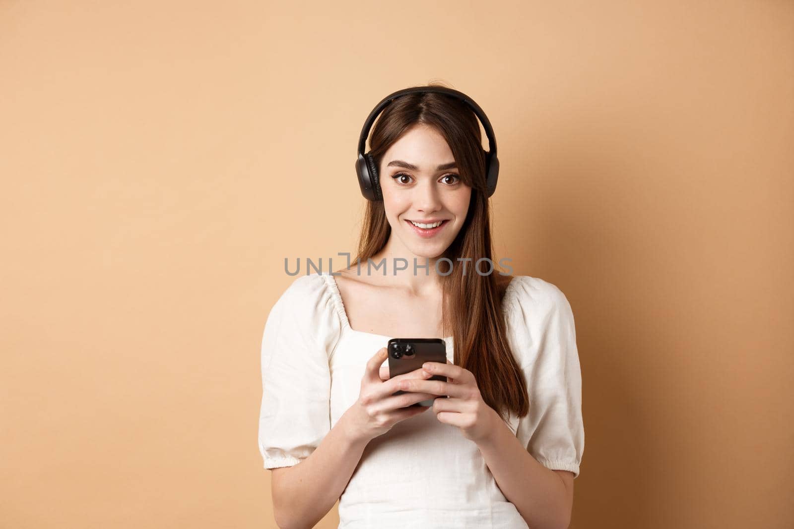 Pretty girl in headphones smiling at camera, listening music and using mobile phone app, beige background.