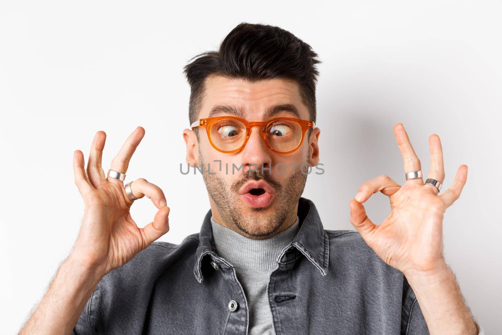 Funny young man in stylish glasses squinting eyes, showing okay gesture, recommend eyewear shop, standing on white background.