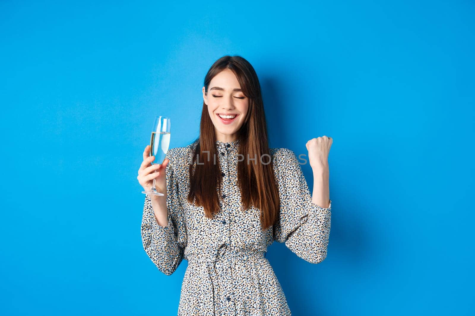Happy woman celebrating win with glass of champagne, dancing satisfiied, triumphing of victory, standing against blue background.