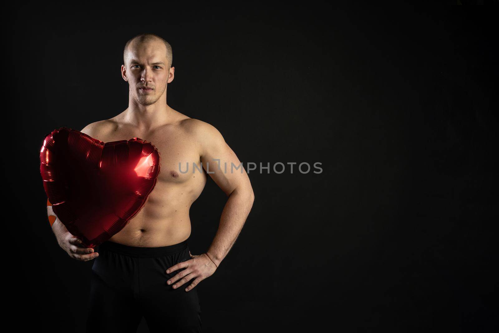 Athletic man holding a red heart, a naked balloon in his hand for a day of velentin pumped up, bodybuilder romantic. lover guy LGBT. In the studio on a black background Cheerful looks at the camera by 89167702191