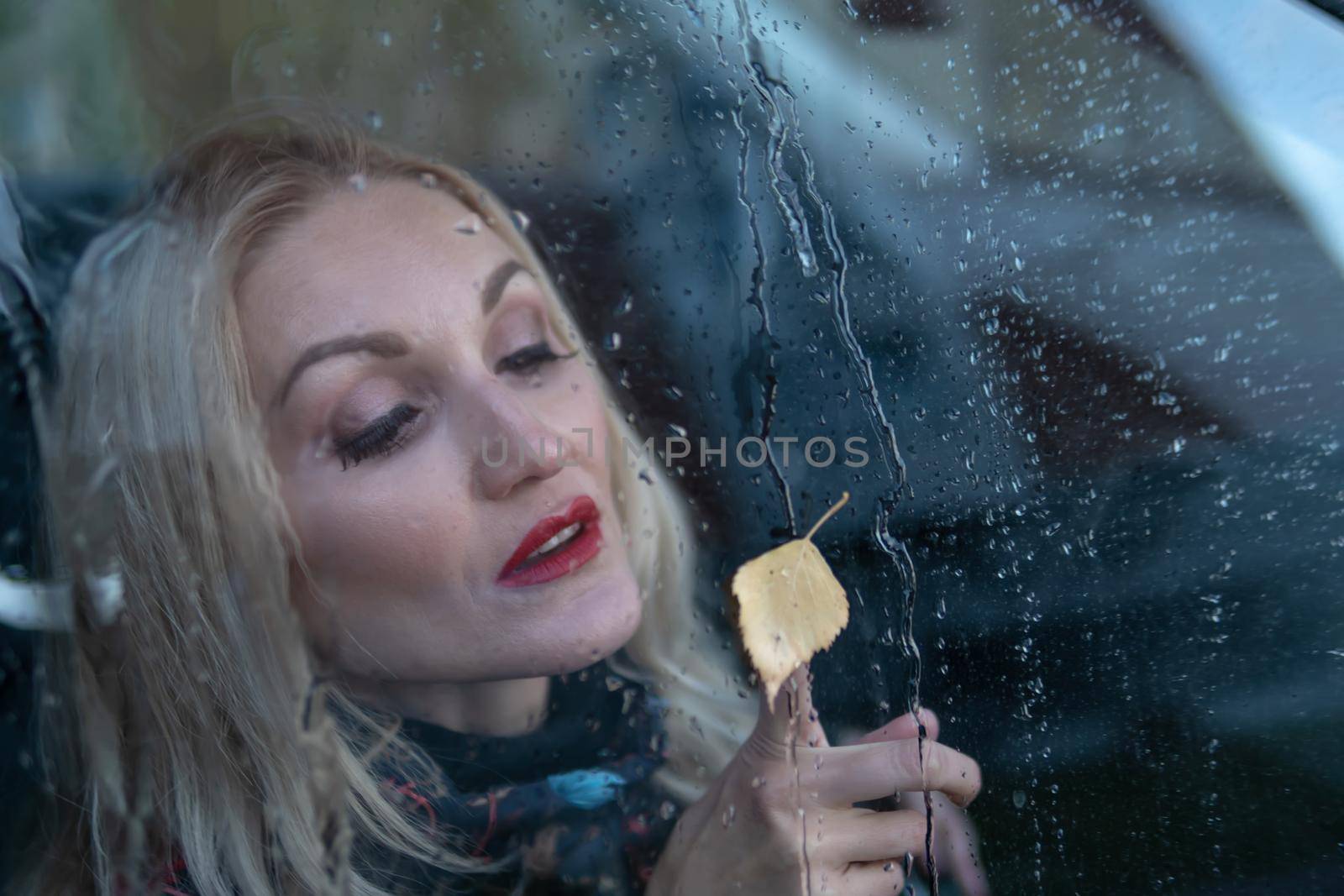 looks through the rainy glass, drops a young girl from the car window on the rain, in a black car. smiling at camera
