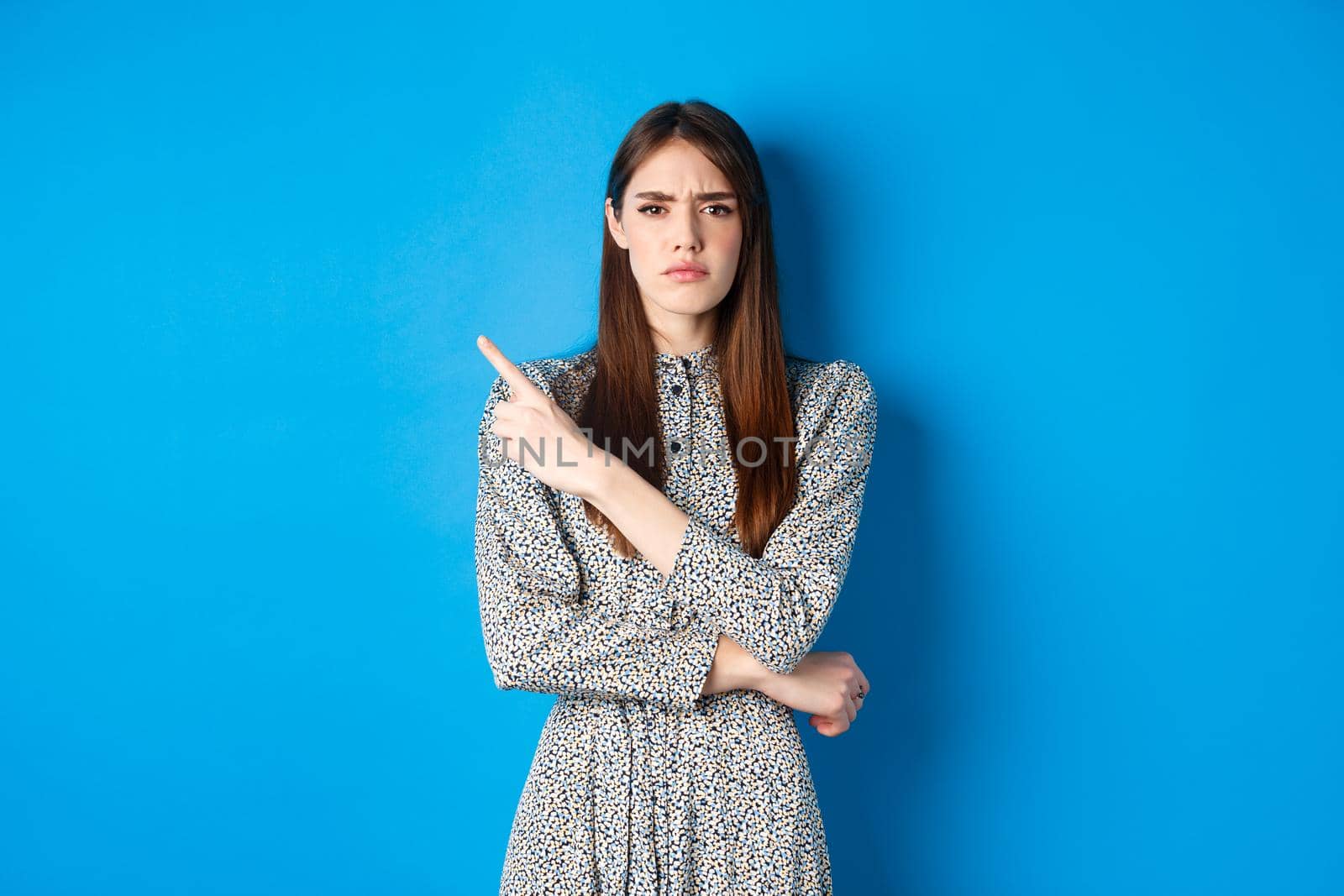 Disappointed frowning girl pointing left and looking displeased, condemn something bad, complaining at banner, standing against blue background in dress.