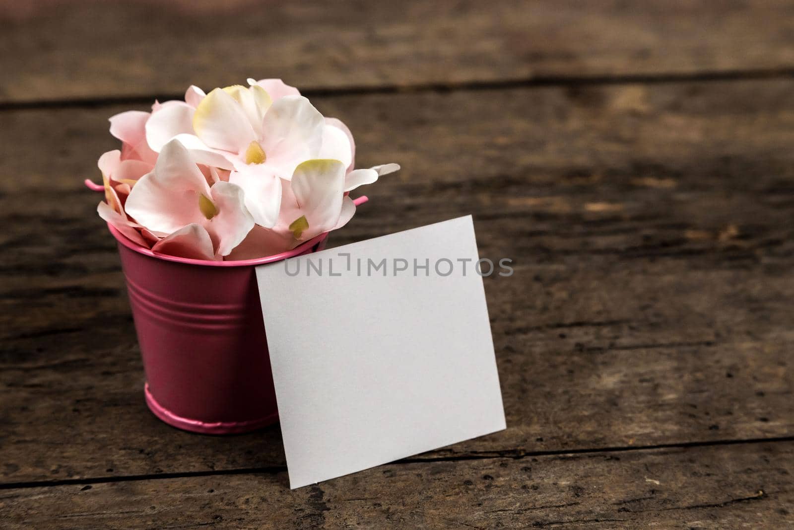 Pastel pink hydrangea flowers in a little bucket and a piece of paper on wood background. Copy space for text.