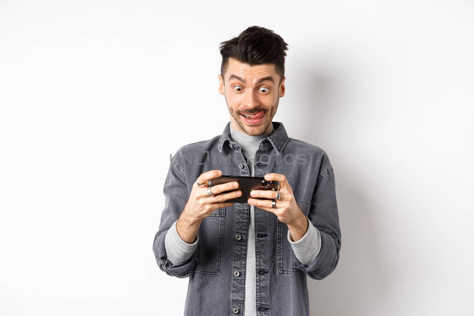 Excited guy playing video game on mobile phone with focused face, tap on smartphone screen, holding cellphone horizontally, white background.