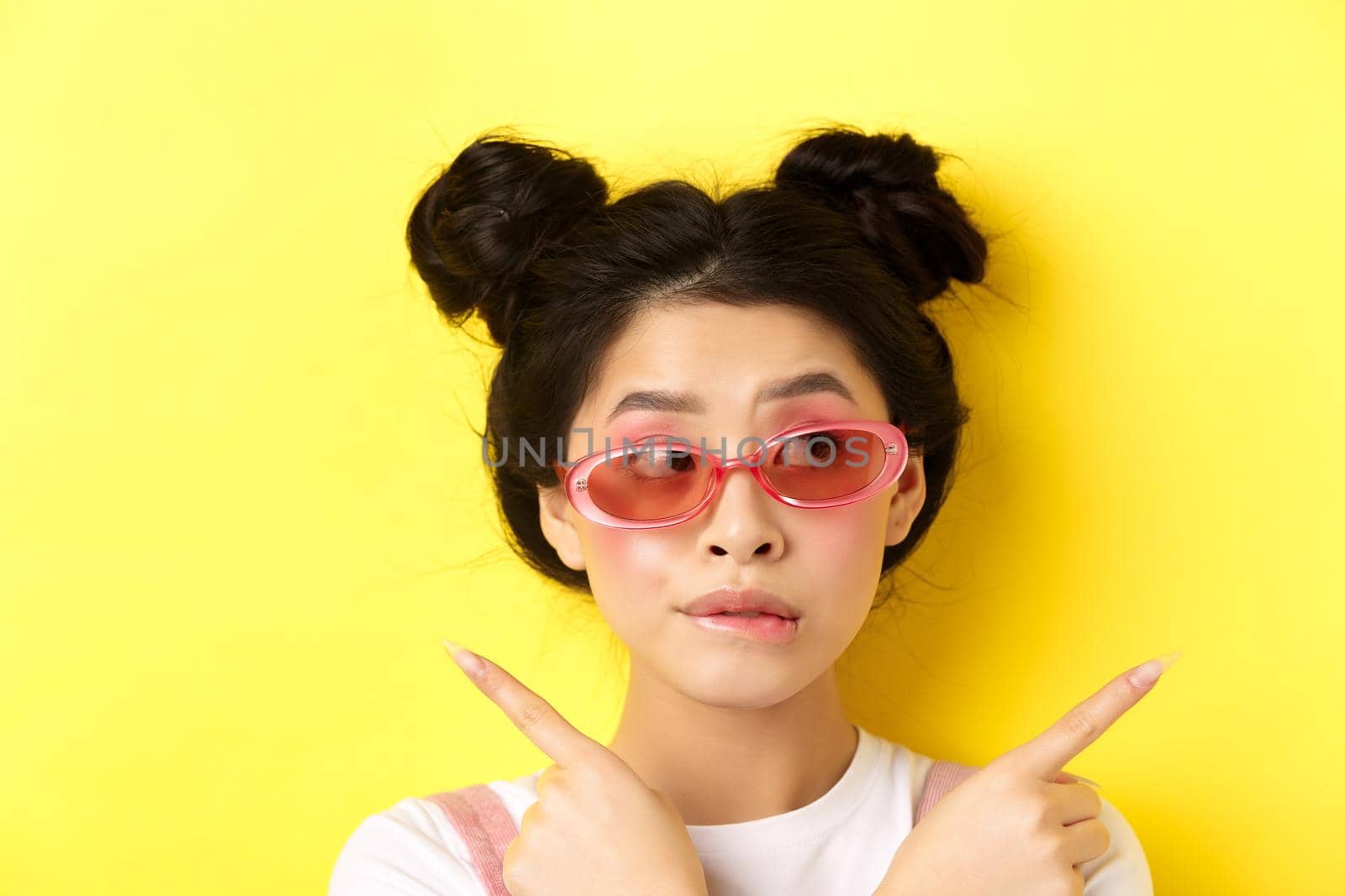 Summer fashion concept. Pensive asian girl making choice, pointing sideways and looking thoughtful, deciding what pick, standing in sunglasses on yellow background by Benzoix