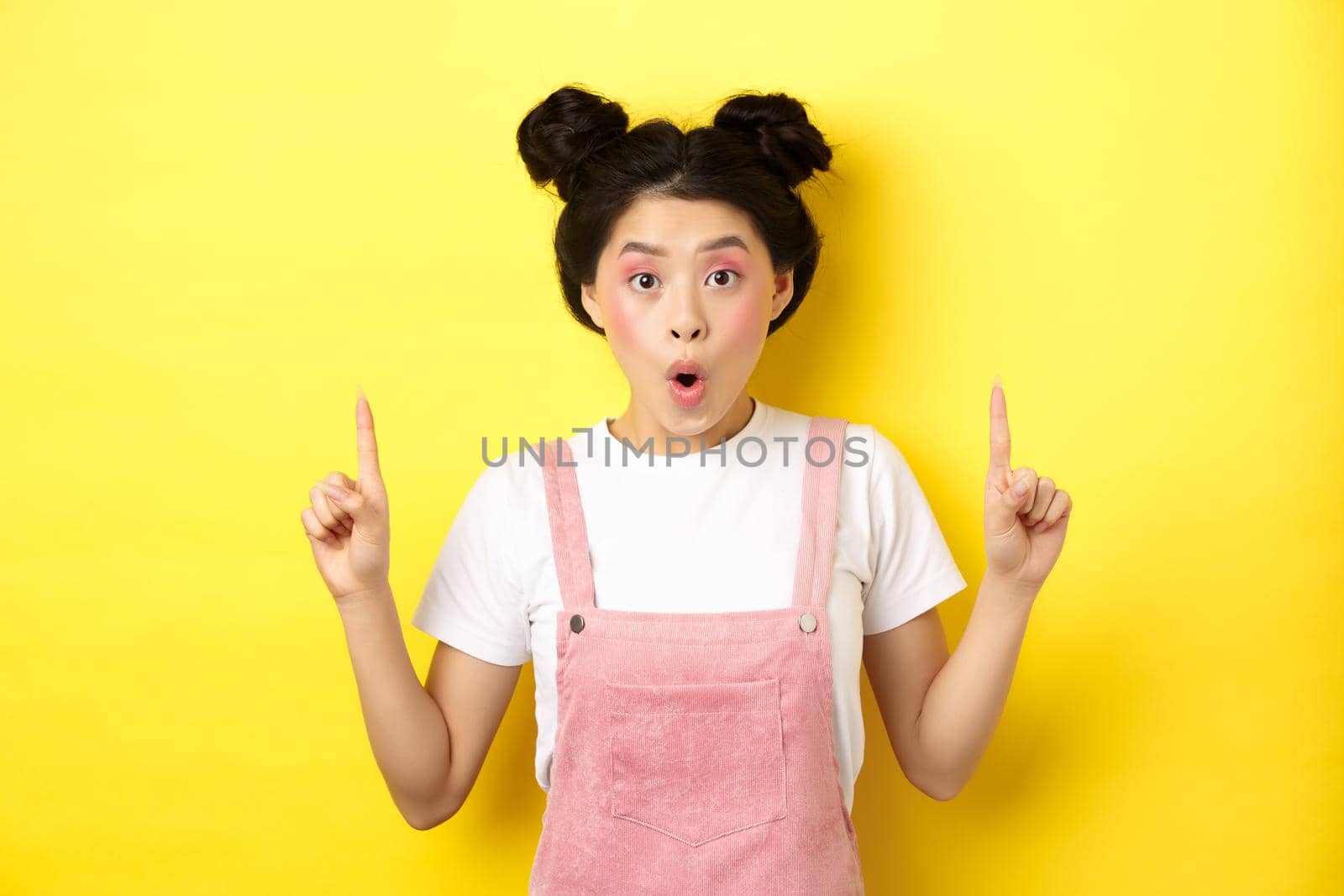 Excited asian female model with glamour makeup, pointing fingers up and say wow amazed, checking out promo deal, standing on yellow background by Benzoix