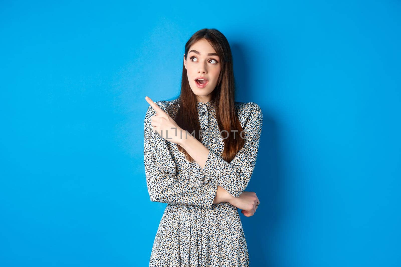 Wow look there. Excited pretty lady in dress looking and pointing left wondered, checking out advertisement, standing on blue background.